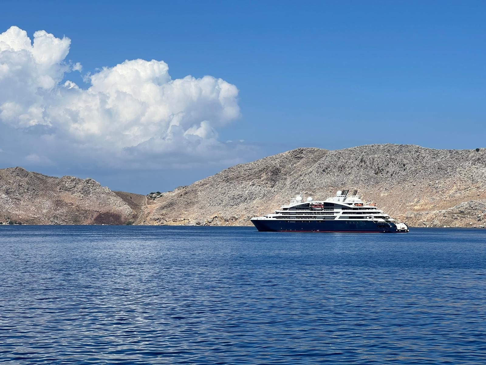 An expedition cruise ship at anchor in a calm blue bay below rocky hills.