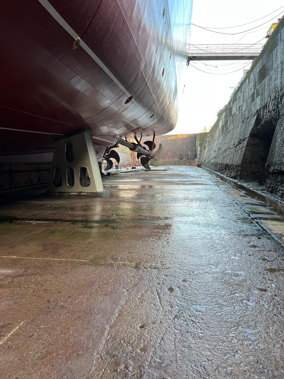 Looking along the floor of a dry dock beneath a ship's hull, with its twin propellers in the distance.