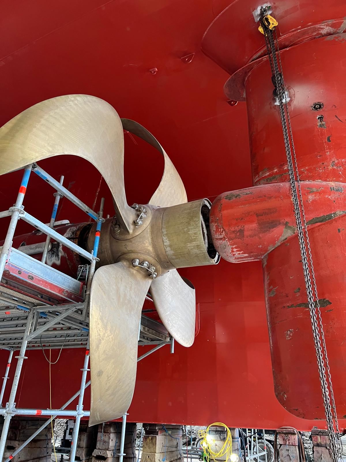 A five-bladed bronze propeller and rudder on a red hull, surrounded by scaffolding in dry dock.